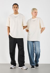 Two individuals wear oversized cream t-shirts with printed logos. One pairs it with black trousers, the other with light blue jeans.