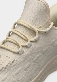 Μπεζ πλεκτό αθλητικό παπούτσι με ελαστικά κορδόνια σε σχήμα βρόχου πάνω από υφασμένη επάνω επιφάνεια και παχύ, διακοσμημένο πέλμα ορατό στην πλευρά.