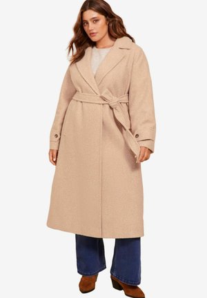 Beige, lang uldblandede frakke med bindebånd, skrå krave og sidelommer. Båret over en lys sweater og parret med udvidede jeans.