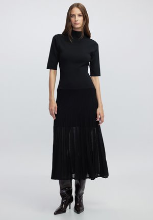 Top noir à manches courtes à côtes associé à une jupe longue plissée noire. Le modèle porte des bottes brillantes jusqu'aux genoux. Tissu lisse, silhouette près du corps.