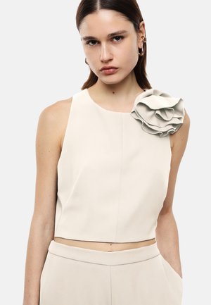 Donna che indossa un top beige senza maniche con un grande fiore di tessuto sulla spalla sinistra e pantaloni coordinati a vita alta, posando con una mano in tasca.