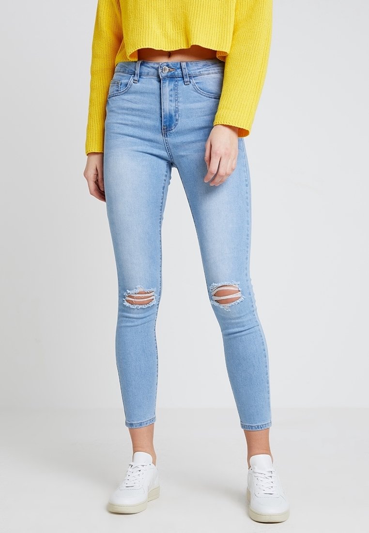 Femme portant un jean skinny déchiré bleu clair, un pull court jaune et des baskets blanches, debout devant un fond clair uni.
