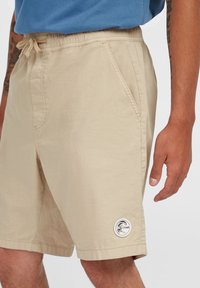 Beige shorts med elastisk midjeband och snörning, med sidofickor och ett runt logopatch på vänster ben. Slät tygtextur.