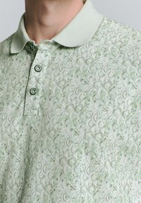 Lichtgroen polo-shirt met een gestructureerd patroon, drie knopen en een zachte kraag. Stof lijkt geweven met subtiele kleurvariaties.