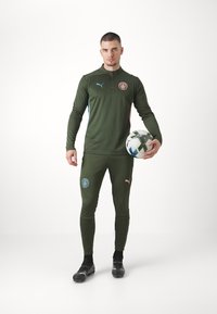 Zöld sportmelegítő vékony szabásban, zipzáras nyakkal és kék akcentussal. Logófoltokkal és texturált anyaggal rendelkezik. Fekete focicipők és labda.
