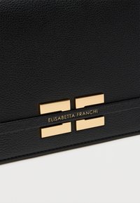 Μαύρη δερμάτινη τσάντα με υφή, διακοσμημένη με λογότυπο σε χρυσό και δύο ορθογώνιες μεταλλικές λεπτομέρειες στο μπροστινό μέρος.