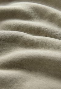 Tekstuuri, hele khaki kangas, millel on nähtav kudumismuster, näidates pehmeid, voolavaid voldikesi ja kerget sära valguse all.