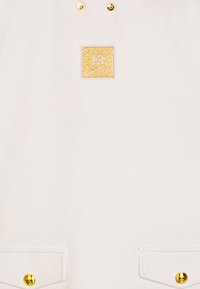 Top in tessuto bianco con etichetta quadrata dorata, due borchie dorate e dettagli con bottoni dorati sulle tasche, dal texture liscia.