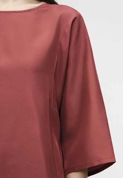 Γυναίκα που φοράει ένα φαρδύ σατέν πουκάμισο σε σκουριασμένο χρώμα με μανίκια μέχρι τους αγκώνες και σκάφος λαιμόκοψη, σε απλό φόντο.