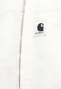 Λευκό ύφασμα με κάθετο μεταλλικό φερμουάρ και ένα μικρό μαύρο και λευκό patch λογότυπο Carhartt ραμμένο κοντά στο φερμουάρ.