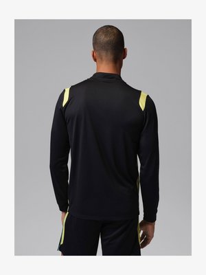 Rövid hajú férfi fekete, hosszú ujjú sportfelsőben és sárga díszítésű rövidnadrágban, hátulnézetben, szürke háttér előtt.