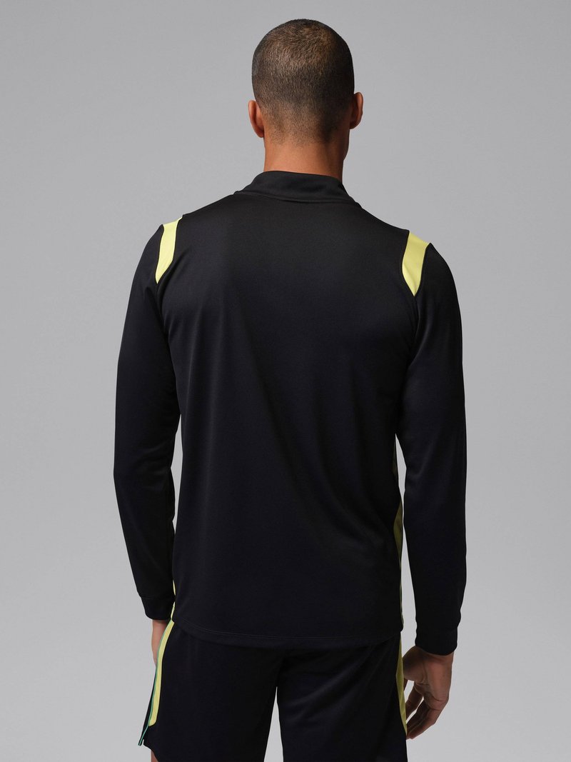 Hombre con cabello corto, vistiendo una camiseta deportiva negra de manga larga y pantalones cortos con detalles amarillos, visto desde atrás sobre un fondo gris.