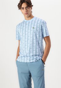 Lichtblauw katoenen t-shirt met een geometrische print in een donkerder blauw. Een klein groen logo is gepositioneerd op de linker borst.