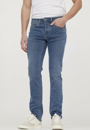 Blue denim jeans with a slim fit, tonal stitching, five pockets, and a zip fly. Worn with white sneakers and a plain white t-shirt.