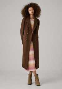 Brown wool overcoat with double-breasted buttons, worn over a striped pastel dress. Beige ankle boots with a structured heel. Textured hair.