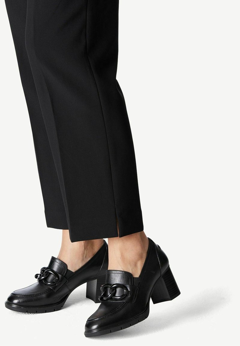 Mocassins en cuir noir à talon bloc avec détail chaîne portés avec un pantalon noir court, vus de la moitié du mollet vers le bas sur fond blanc.
