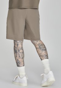 Helle braune Shorts mit einer glatten Textur, kombiniert mit weißen Turnschuhen und gerippten weißen Socken. Detaillierte Tattoos sind an den Beinen sichtbar.