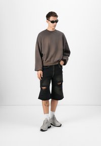 Sweatshirt cinza oversized, calções de denim preto desgastados com destacados rasgos, ténis cinzentos e óculos de sol escuros. Fundo limpo.