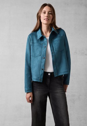 Giacca in finta suede color verde acqua con colletto piccolo, due tasche sul petto e chiusure a pulsante. Abbinata a jeans neri a gamba larga.