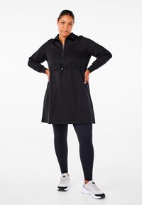 Melnā zīmolējamā kleita ar pieguļošu jostasvieta un sānu kabatām, papildināta ar tumši zilu legingu un pelēkiem sporta apaviem. Gluda auduma faktūra.