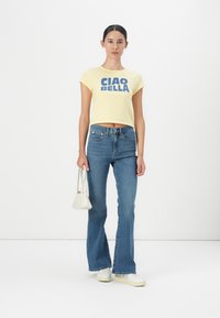 Yellow cropped t-shirt with blue "CIAO BELLA" text, paired with flared blue jeans and white sneakers, holding a small white handbag.