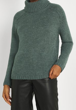 Personne portant un pull à col roulé vert en tricot et un pantalon en cuir noir, debout avec une main dans la poche et l'autre détendue.