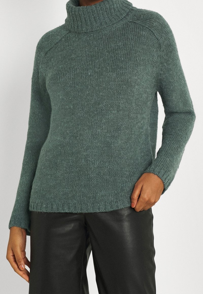 Personne portant un pull à col roulé vert en tricot et un pantalon en cuir noir, debout avec une main dans la poche et l'autre détendue.