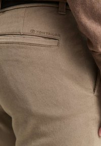 Gros plan sur un pantalon beige texturé avec une poche arrière et un logo "Tom Tailor" embossé au-dessus de la poche.