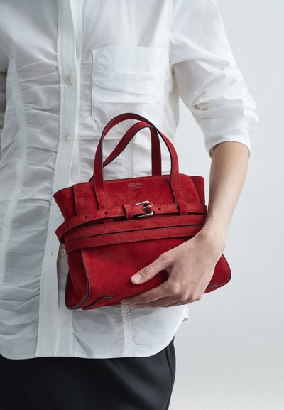 Person som bär en vit rynkad skjorta och svart kjol, hållande en liten röd mockaväskan med spännband.