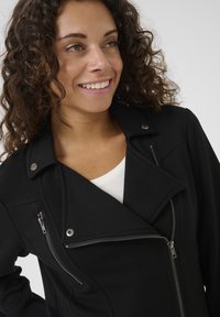 Svart jacka med asymmetrisk dragkedja, strukturerad tyg, detaljer i silver och slagkrage. Bärs över en vit topp.