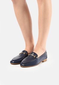 Mocasines de cuero azul marino con cadena decorativa en tono dorado, punta redonda, tacón bajo y textura suave, combinados con piernas al aire.