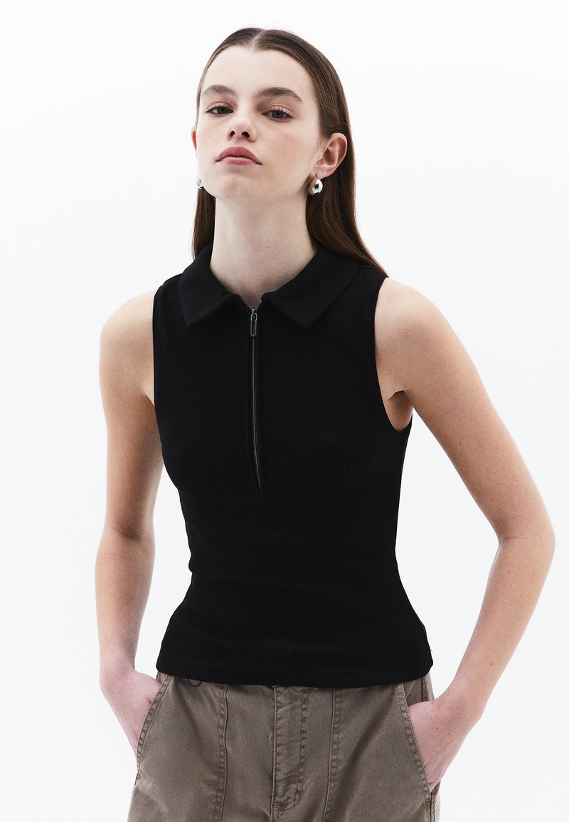 Top sans manches noir avec une fermeture éclair et un col. Présente un design ajusté et une texture côtelée. Le modèle l'associe avec un pantalon marron clair.