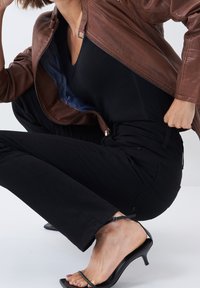 Personne accroupie portant une veste en cuir marron, un top noir, un pantalon noir et des sandales à talons noires avec des brides à la cheville.