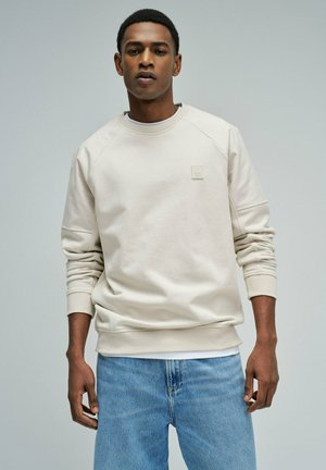 Joven con sudadera beige clara y jeans azules, de pie frente a un fondo gris liso, mirando hacia adelante.