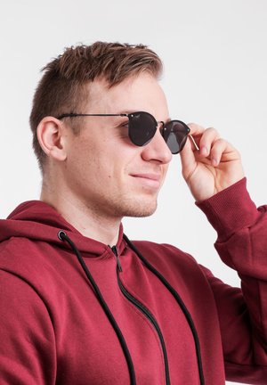 Kulaté černé sluneční brýle s tenkým kovovým rámem, hladkými skly a decentním brandingem. Model má na sobě vínovou mikinu na zip.