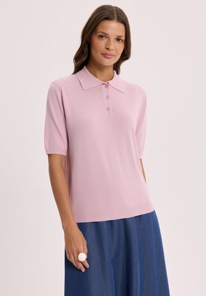 Kobieta ubrana w jasnoróżową koszulkę polo z krótkim rękawem i niebieską spódnicę, stojąca z neutralnym wyrazem twarzy na jasnym tle.