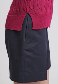 Vue de côté d'une personne portant un short bleu marine foncé avec poche fendue et un pull rouge tricoté avec des fentes latérales.