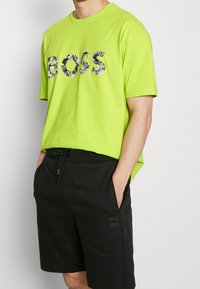 Světlá limetková bavlněná trička s logem v maskáčovém vzoru, které hlásá "BOSS." V páru s černými šortkami s přítlačným logem.