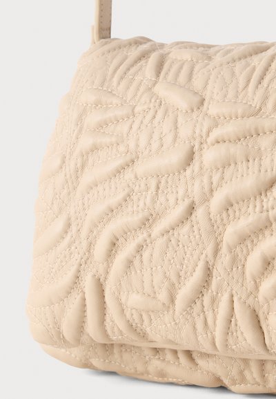 Μπεζ δερμάτινη τσάντα με επένδυση σε πουά μοτίβα με ανυψωμένες, ακανόνιστα ραμμένες ραφές και λεπτό ιμάντα ορατό στην κορυφή.