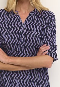 Knopfzyklusbluse in Marineblau mit lila Wellenmuster, ausgestattet mit einem entspannten Kragen und hochgekrempelten Kurzärmeln. Glatte Stofftextur.