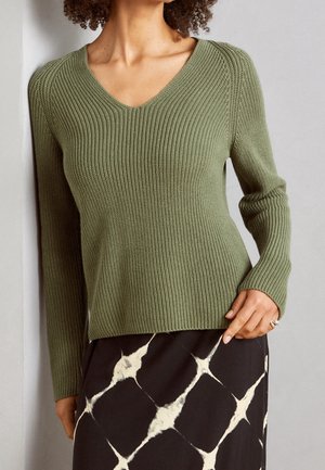 Un pull en maille côtelée vert olive avec un col en V et des manches longues, associé à une jupe noire à motifs avec des formes de losanges blancs.