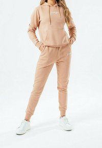 Světle béžová mikina a odpovídající tepláky vyrobené z měkké tkaniny, s přední kapsou, detaily na šňůrkách a bílými logovými akcenty. Doplněno bílými teniskami.