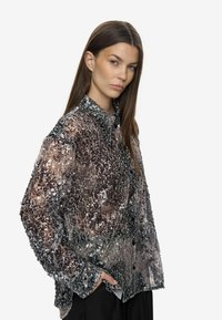 Femme aux longs cheveux bruns portant une blouse noire et argentée à manches longues et à sequins, posant avec une main dans la poche.