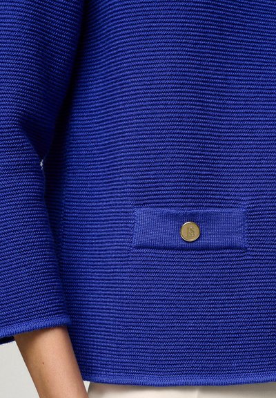 Blaues Strickpullover mit einem strukturierten Muster, ausgestattet mit einer kleinen Tasche und einem runden Metall-Logo-Knopf auf der Vorderseite.