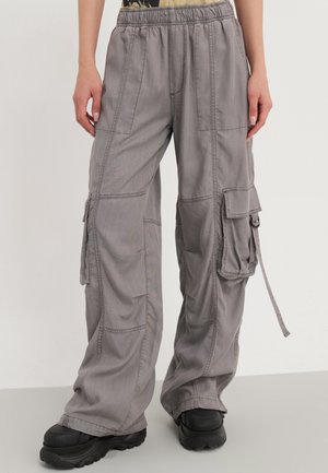 Pantalones cargo grises sueltos con múltiples bolsillos y detalle de cordón, usados con zapatos negros gruesos y una vista parcial de las manos.