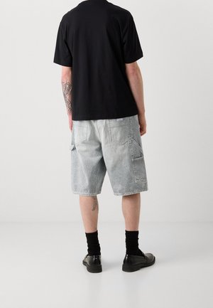 Man draagt een zwart T-shirt met korte mouwen, lichtgrijze cargo shorts tot over de knieën, zwarte sokken en zwarte schoenen, staat met zijn rug naar voren op een witte vloer.