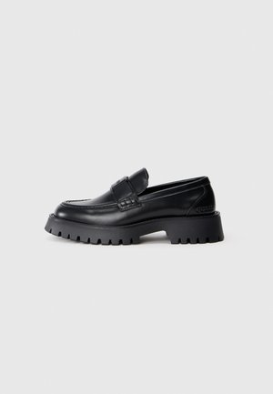 Mocassin en cuir noir avec une semelle épaisse à crampons et une bride sur le dessus, vue de côté sur fond gris clair uni.