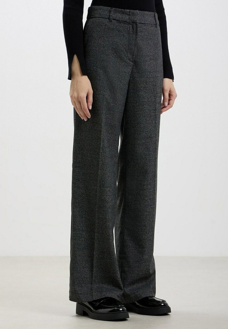 Pantalons larges en tissu texturé gris foncé, dotés d'une taille haute, de passants de ceinture et d'un motif à carreaux subtil. Mocassins noirs brillants.