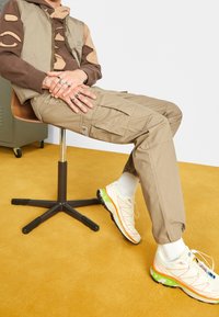 Beiget työhaalarihousut sivutaskuilla, ruskea korjattu huppari ja valkoiset lenkkarit, joissa värikkäitä yksityiskohtia. Istuu puisella tuolilla.