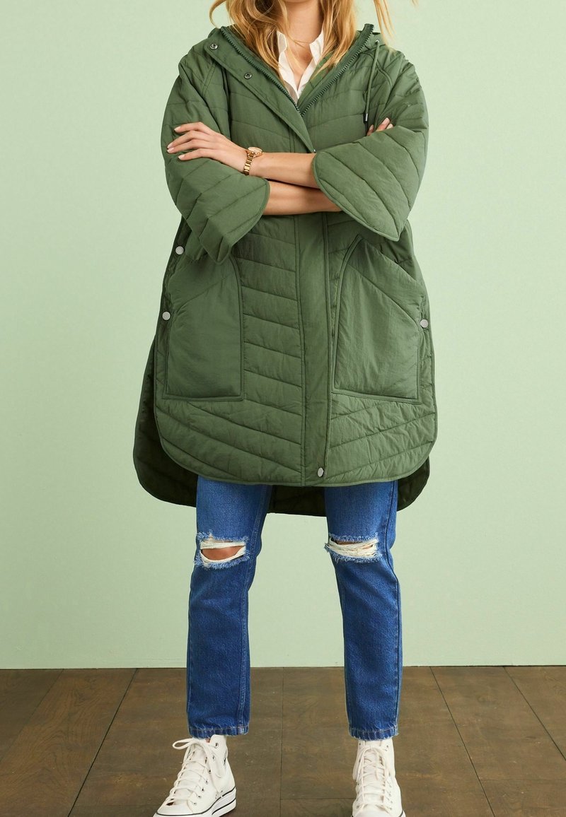 Personne portant une veste matelassée verte avec de grandes poches, un jean bleu déchiré, des baskets blanches et une montre-bracelet en or, debout sur un sol en bois.